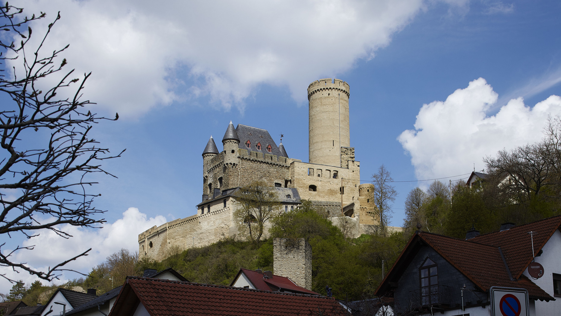Ansicht der Burg Schwalbach, Berfired rechts vor blauem Himmel mit Wolken, links im Vordergrund und rechts Bäume, ein Wohnhaus.